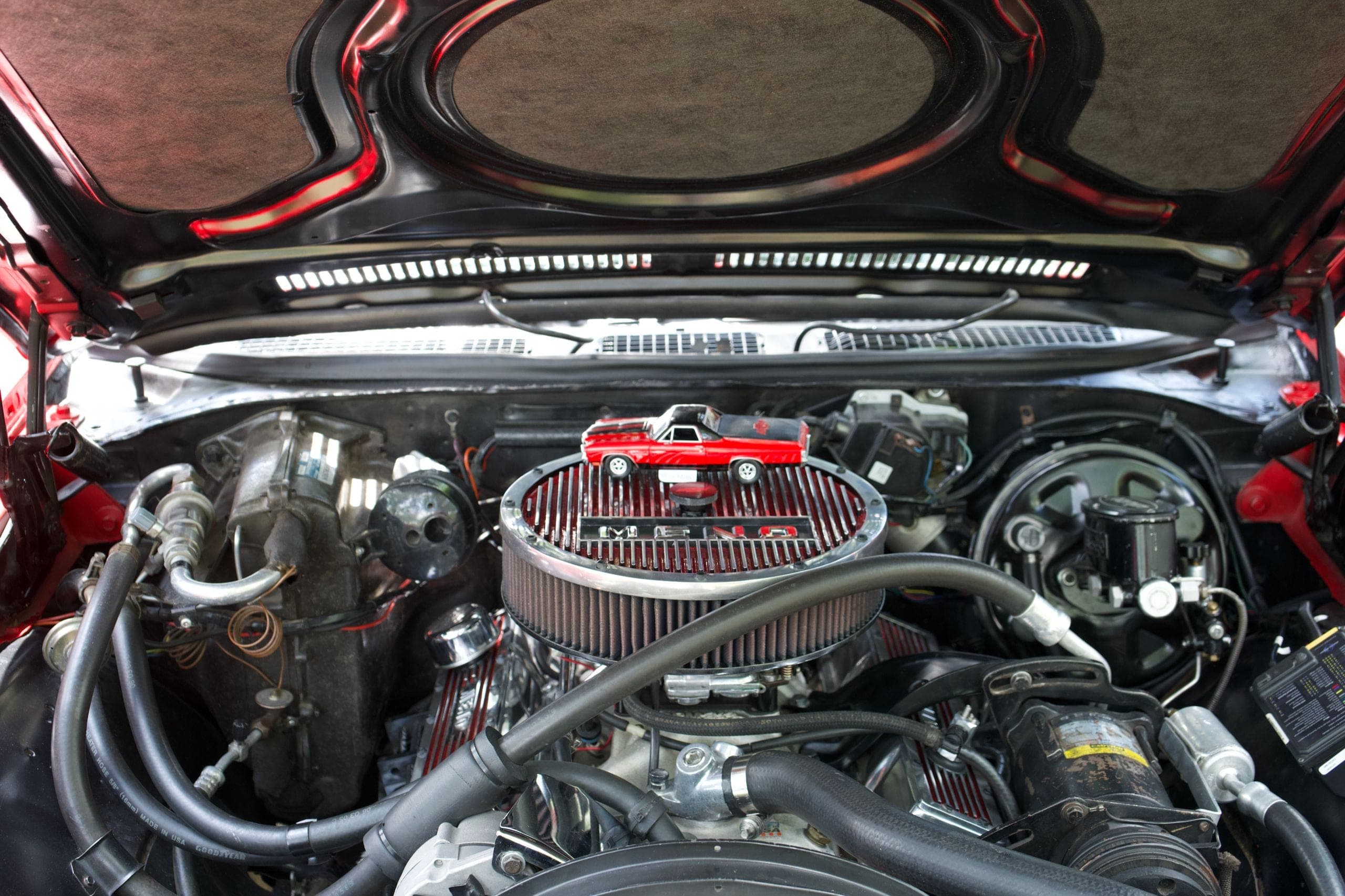 Open engine bay of a red V8 car with a chrome-topped round air cleaner and black rubber hoses, with a small red die-cast pickup set on top of the air cleaner