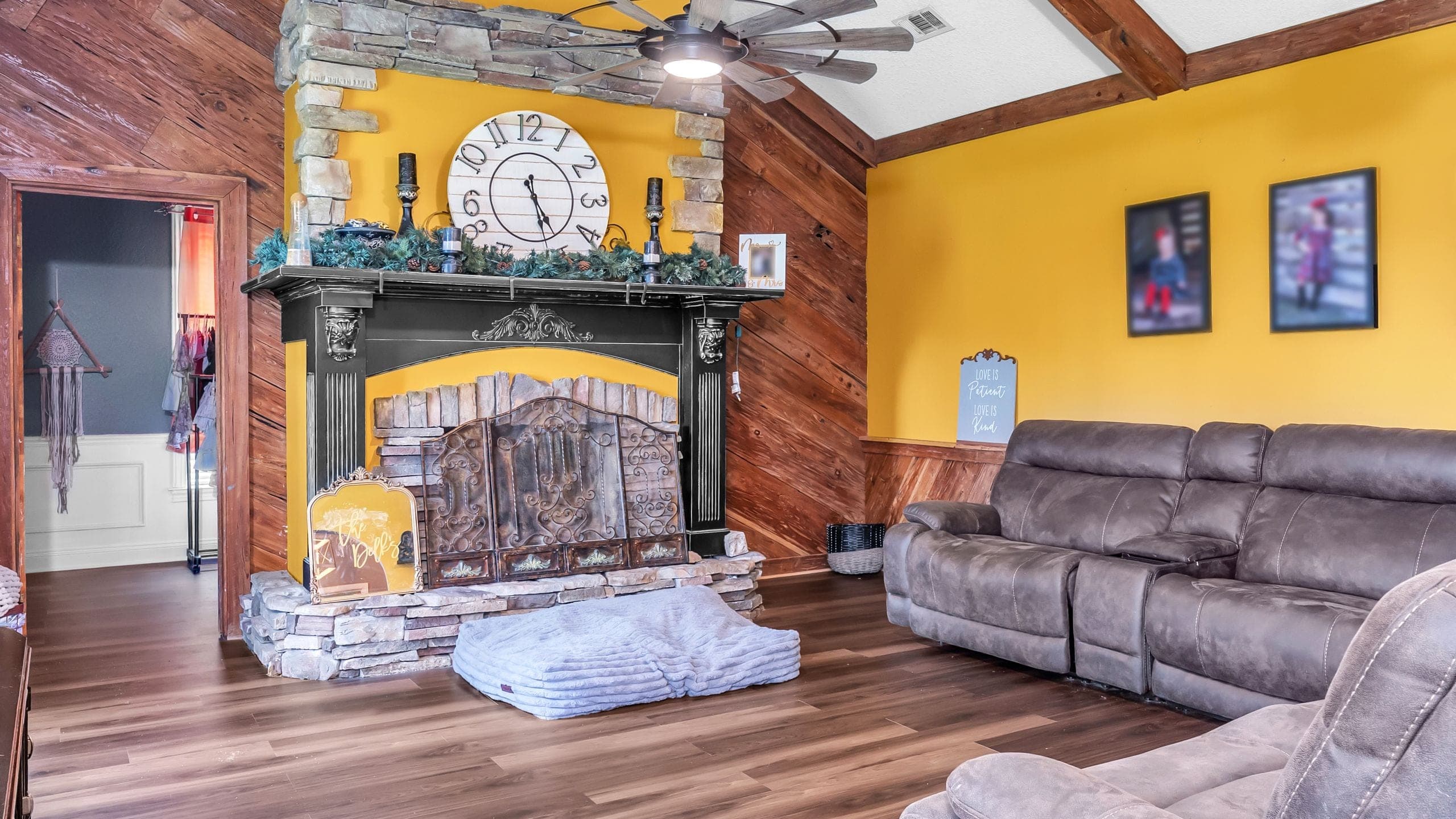 Living room with a stone and black-mantel fireplace against a saturated yellow wall, reclaimed wood panelling and a grey sectional, lit evenly throughout (Flambient edit)