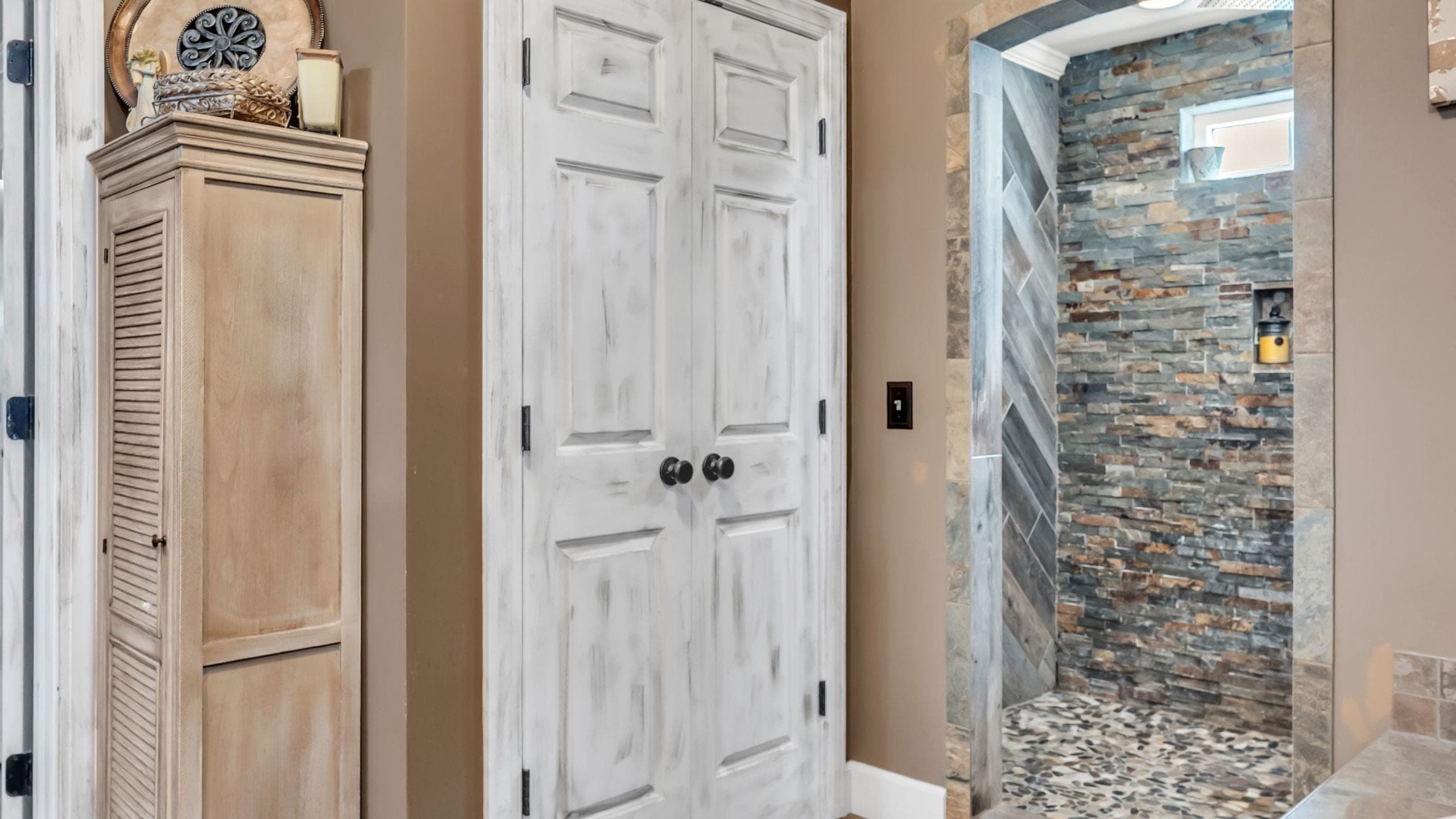 Walk-in shower lined in stacked slate and river-rock tile, seen through a doorway beside a whitewashed linen cabinet (Flambient edit)