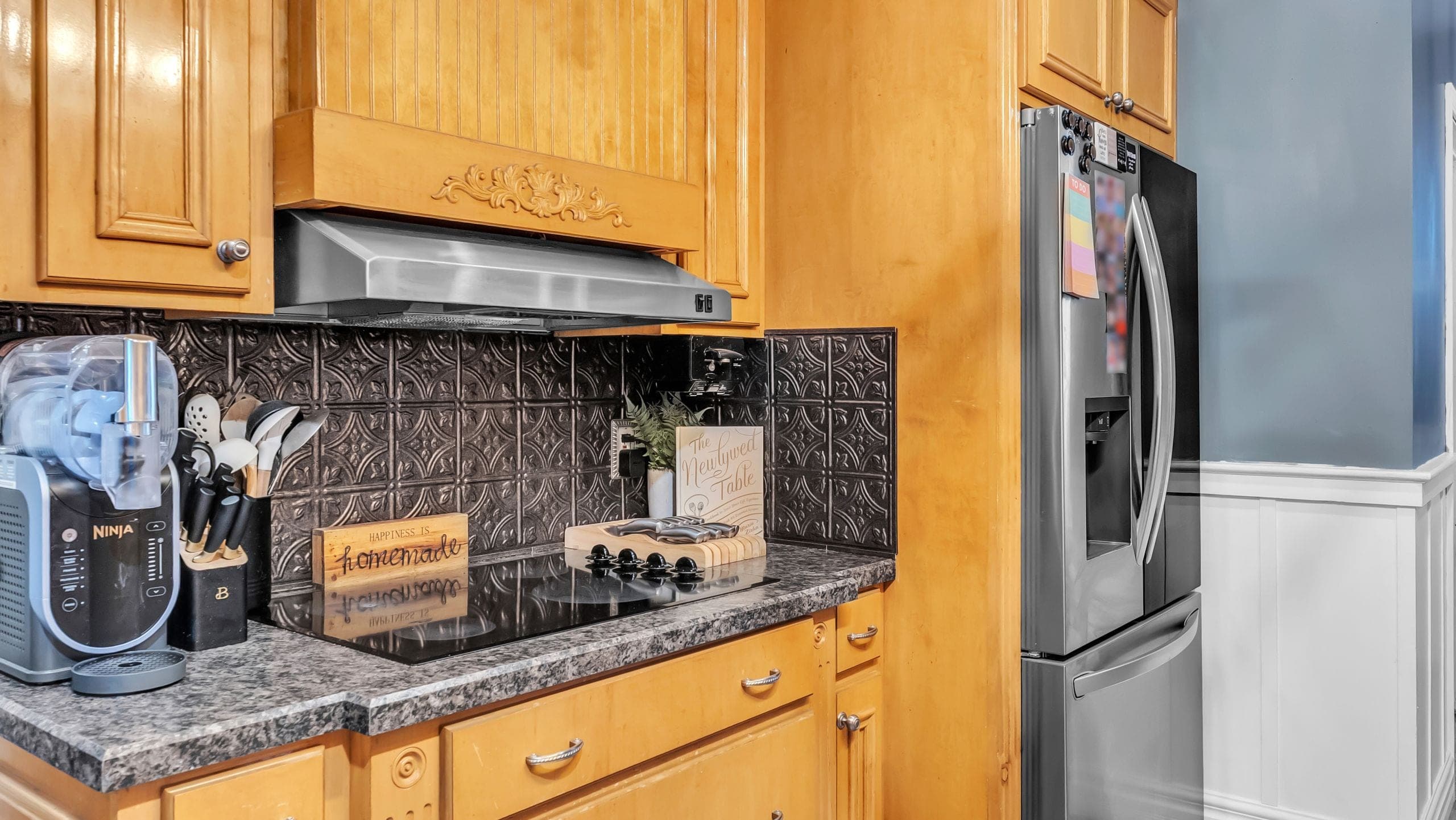 Kitchen with honey oak cabinets, a pressed-tin backsplash and a stainless refrigerator, the dark backsplash keeping its detail against the bright countertop (Flambient edit)