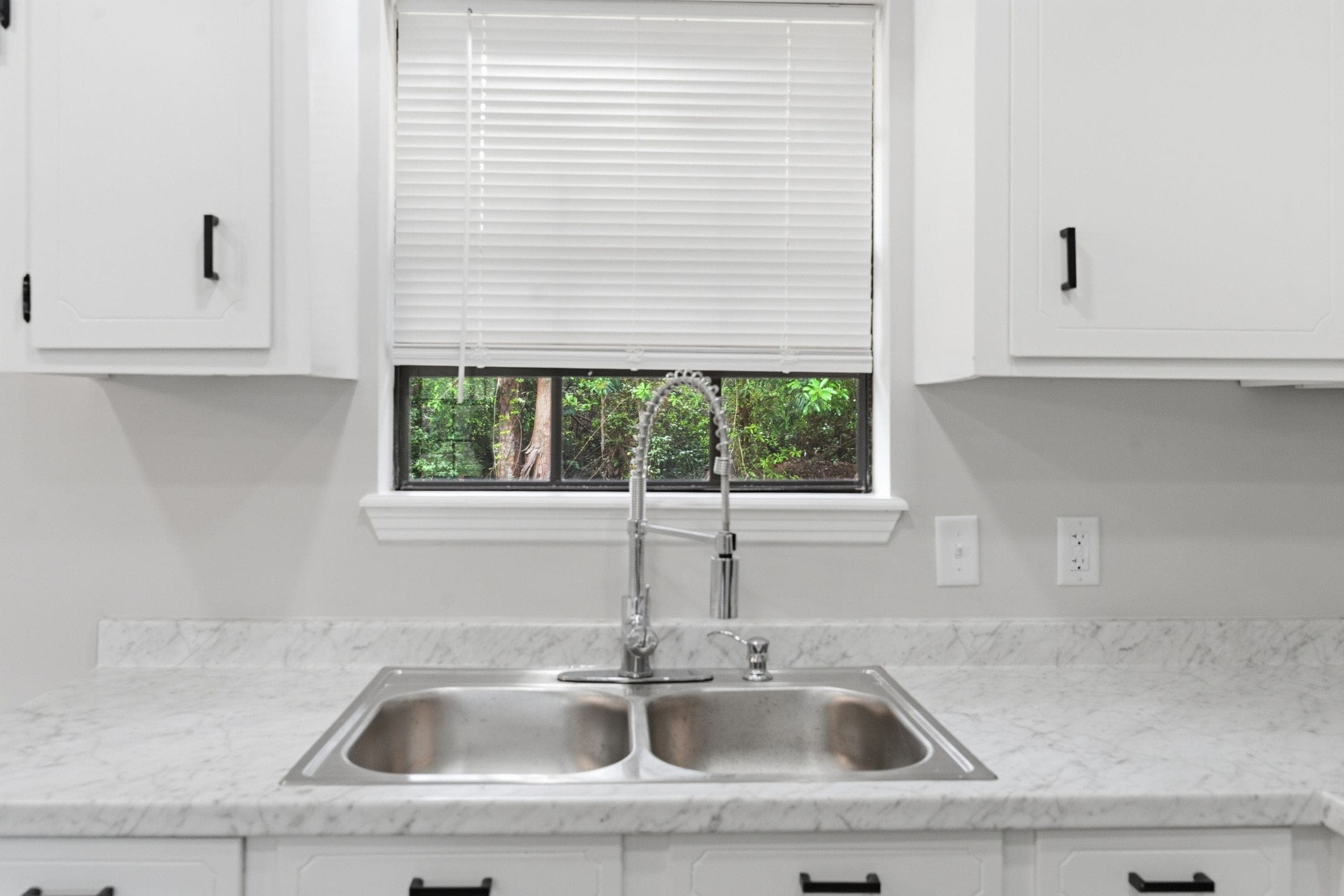 Kitchen sink and pull-down faucet below a window with the blind half raised, the trees outside holding detail instead of blowing out to white (HDR edit)