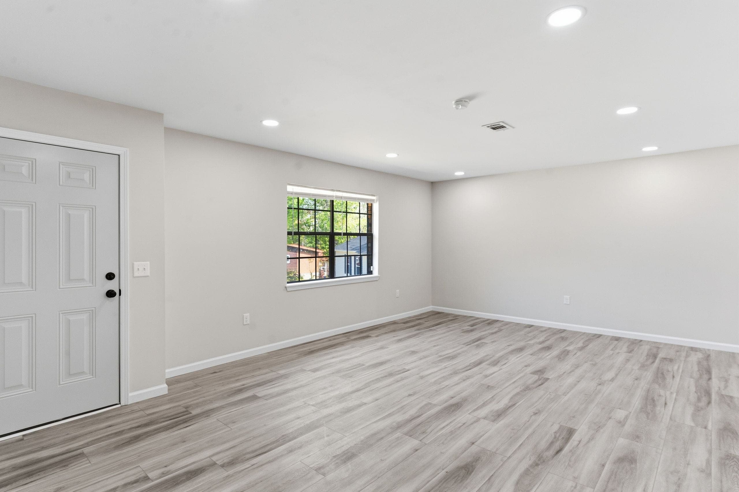 Empty living room with pale plank flooring, a white entry door and a single window showing the yard beyond (HDR edit)