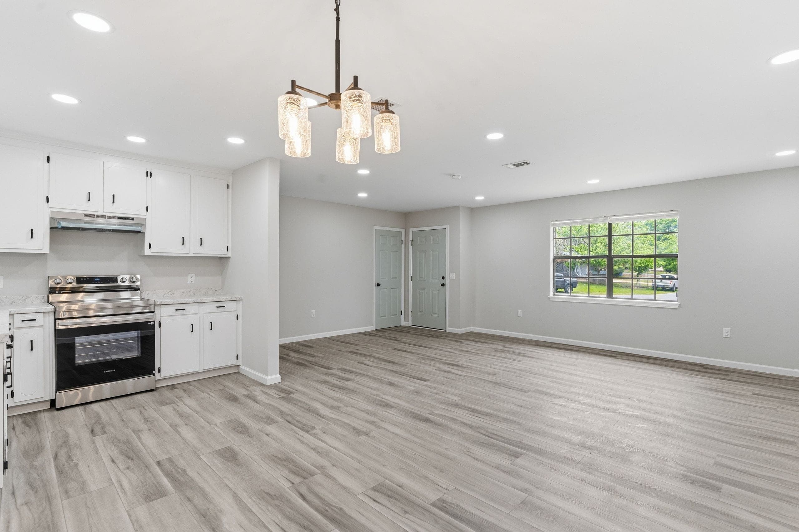 Vacant open-plan kitchen and living area with white shaker cabinets, a stainless range and a glass-globe chandelier, with the view through the far window still legible (HDR edit)