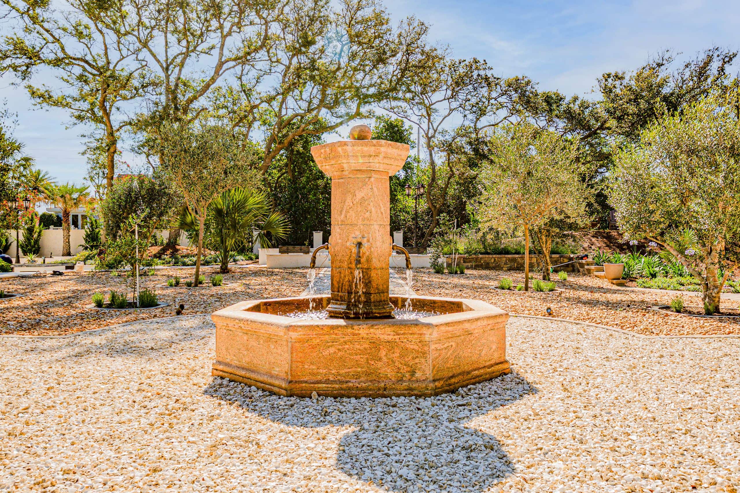 Carved stone fountain running in a crushed-shell courtyard framed by silver-leaved trees and young plantings