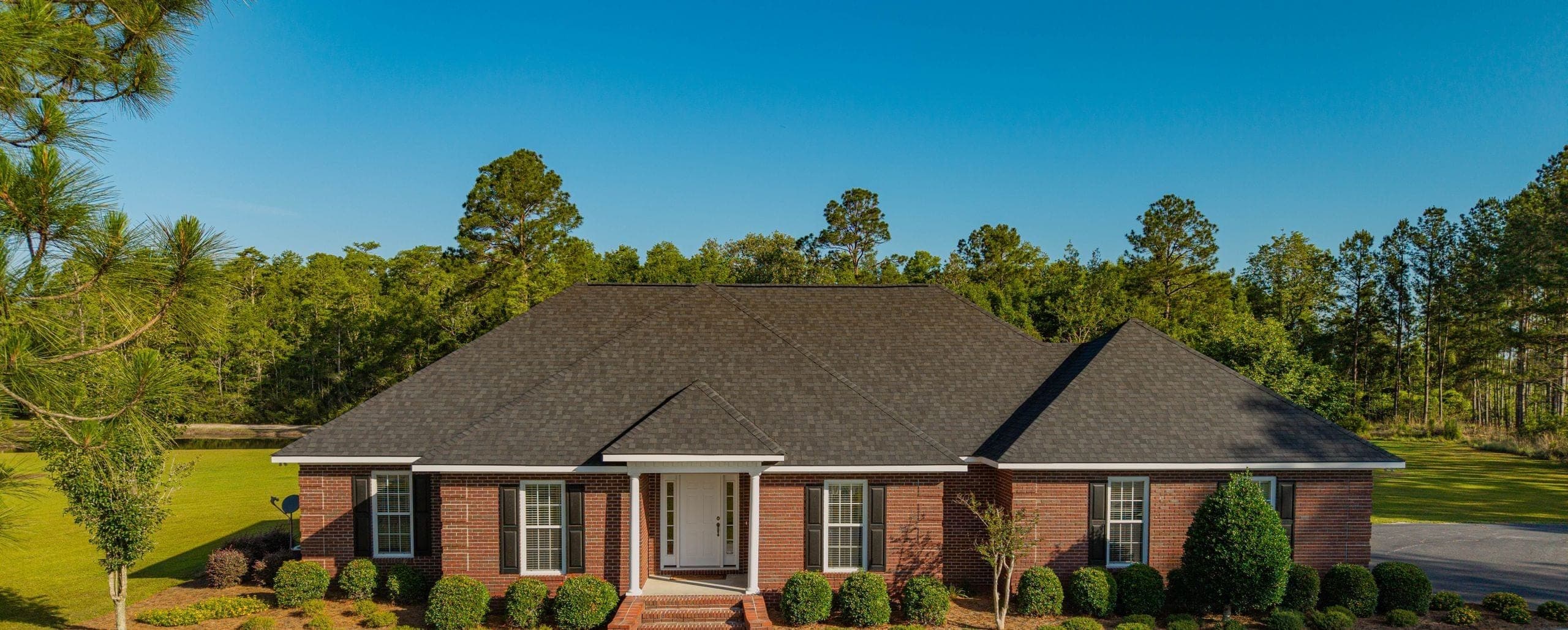 Aerial view of a red brick ranch home with trimmed shrubs and a mown lawn, backed by a pine treeline under a clear sky