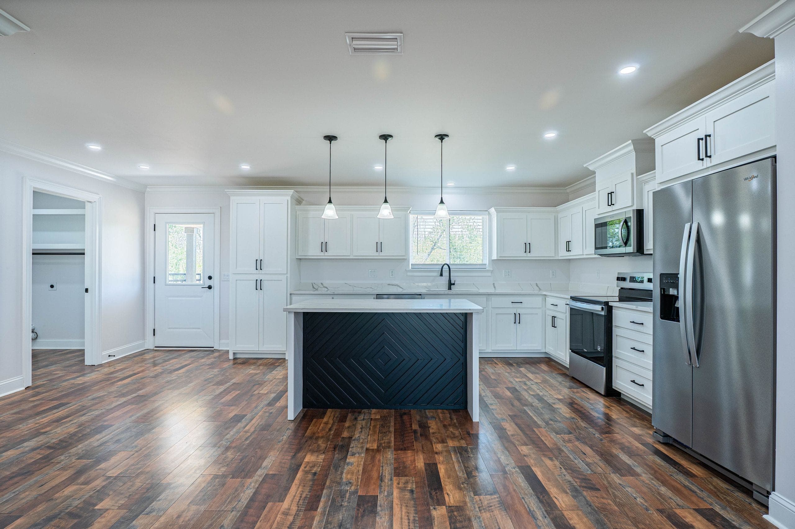 Open kitchen with white shaker cabinets, a black island front in a mitered diamond pattern, three pendant lights and stainless appliances over wide-plank wood-look flooring