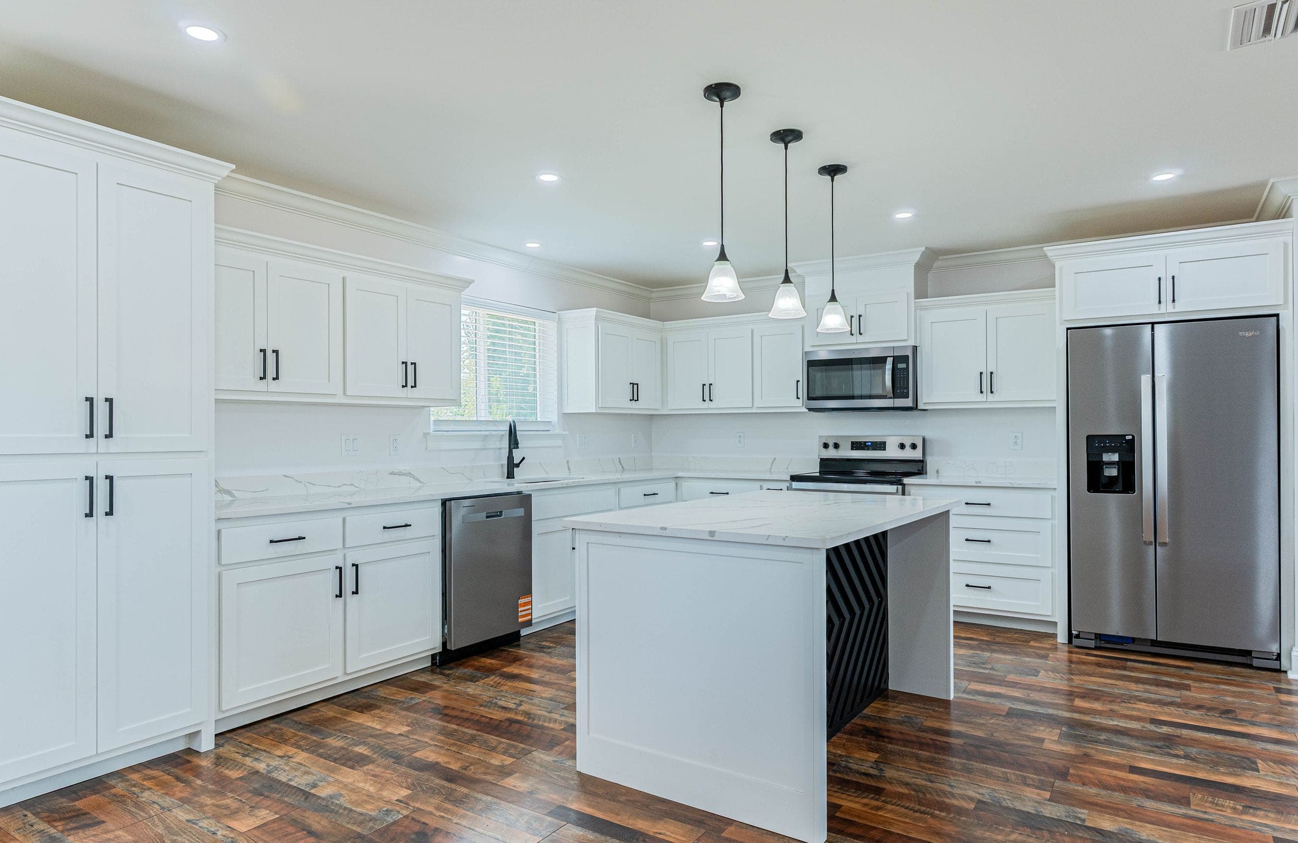Wide view of a white kitchen with floor-to-ceiling cabinetry, marble-veined stone counters, a center island and stainless refrigerator, lit by recessed and pendant lighting