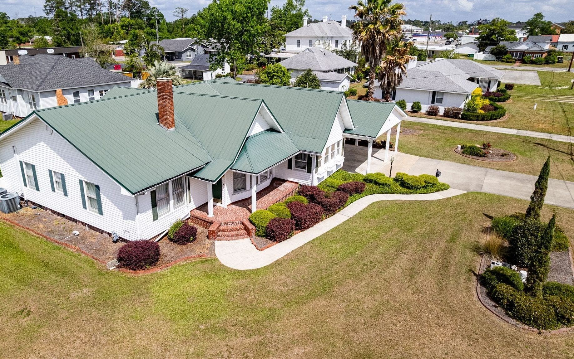 Aerial view of a white farmhouse with a green metal roof, brick chimney and curved front walk, with palms and neighboring houses beyond