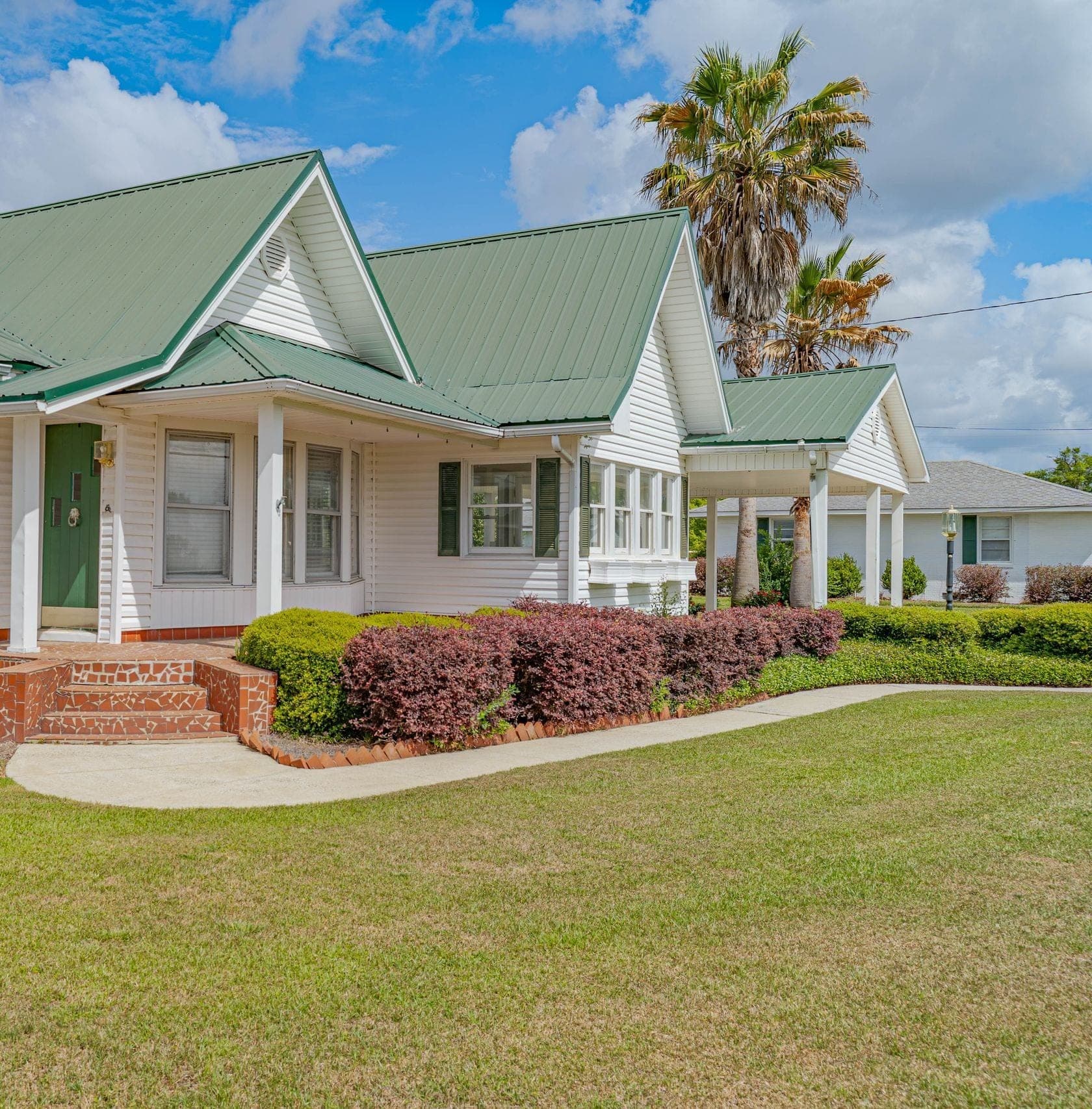 Ground-level view of a white farmhouse with a green metal roof, green shutters, brick steps and a covered porch behind a landscaped front bed