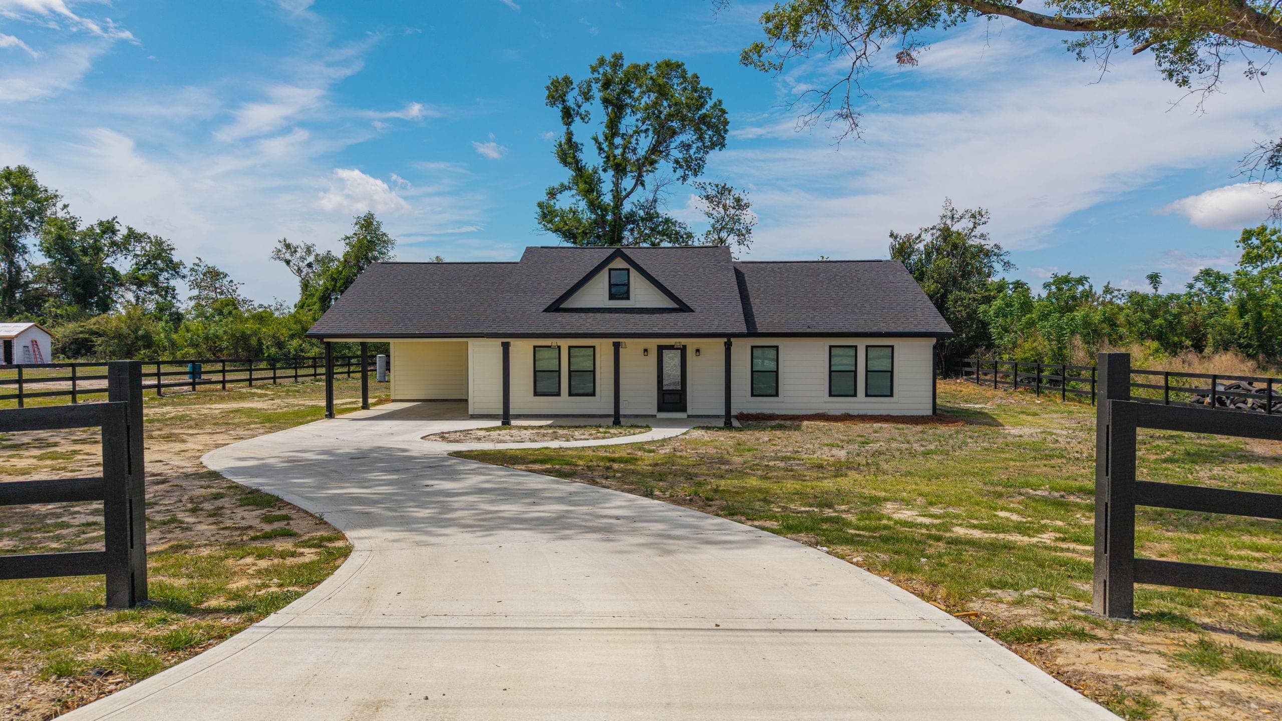 Ground-level view of a cream ranch home with a dark shingled gable roof and attached carport, at the end of a wide concrete driveway between black board fencing